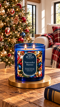 Load image into Gallery viewer, “Lit blue glass candle labeled Mill POND Candle Co., Nutcracker Matinee, Blended Molasses, Candied Almonds, Lemon Zest, sitting on a gold base in front of a decorated Christmas tree with ornaments, nutcracker figures, and a cozy living room scene featuring a white armchair with a red plaid blanket and natural light from a nearby window.”
