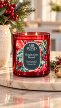 Load image into Gallery viewer, “Red glass candle labeled Sleighcation Bound with two lit wicks sitting on a marble surface, surrounded by festive greenery, red berries, and a gold ornament, with soft holiday décor in the background.”
