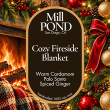 Load image into Gallery viewer, “Brown candle label reading Cozy Fireside Blanket, surrounded by holiday greenery, red ornaments, gold baubles, pinecones, and a red bow with a fireplace in the background.”

