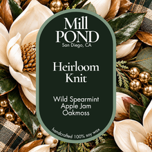 Load image into Gallery viewer, “Green candle label reading Heirloom Knit, surrounded by magnolia flowers, pinecones, green leaves, and gold ornaments.”
