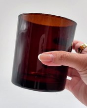 Load image into Gallery viewer, A hand holding a dark amber brown translucent glass tumbler against a plain white background.

