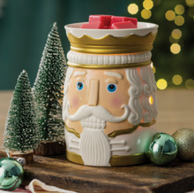 Load image into Gallery viewer, Ceramic nutcracker‑shaped wax warmer sitting on a wooden surface. The warmer features a gold crown, blue eyes, and a white mustache, with red wax cubes placed on top. It is surrounded by small snowy evergreen trees and green ornaments. Warm, blurred holiday lights glow in the background, creating a festive Christmas scene.
